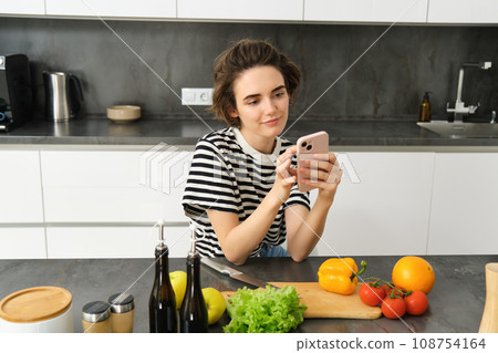 Portrait of cute brunette woman in the kitchen, cooking, searching recipe on smartphone, taking pictures for her food blog, cooking meal from vegetables, vegan salad 108754164