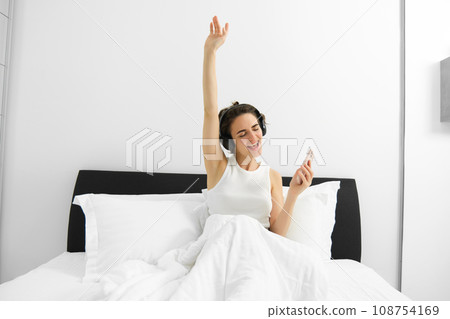 Portrait of excited, happy woman dancing and singing in bed, waking up and listening to music in wireless headphones 108754169