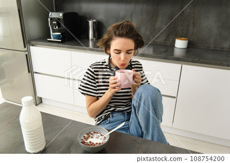 Cute brunette woman, student eating breakfast in morning, drinking hot tea, smiling and looking pleased Cute brunette woman, student eating breakfast in morning, drinking hot tea, smiling and looking pleased 108754200