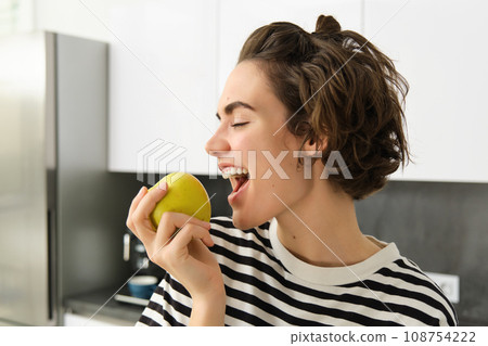Close up portrait of young brunette woman biting an apple with pleasure, has pleased smile on her face, standing in the kitchen, having healthy snack for lunch, eating fruits 108754222