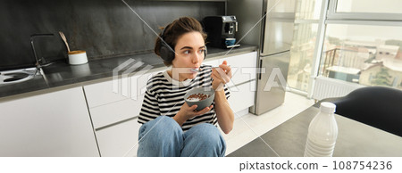 Image of stylish, modern woman, student eating cereals with milk for breakfast, holding bowl and spoon, listening music in wireless black headphones 108754236