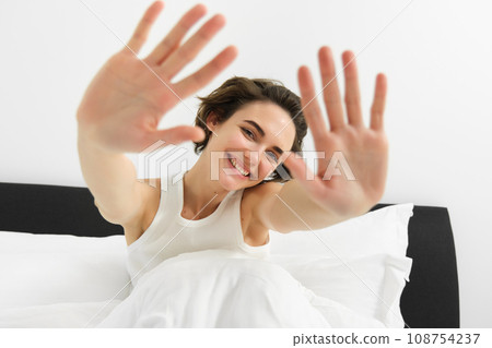 Portrait of sleepy, happy young woman, wakes up in her bed, stretching out hands, hiding face from camera, smiling and laughing 108754237