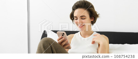Beautiful woman lying in bed with smartphone, resting in her hotel suite, using mobile phone for food delivery, reading on cellphone 108754238