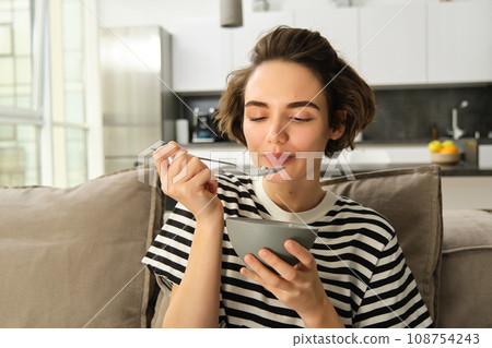 Portrait of young woman eating granola, bowl of cereals with milk, sitting on sofa and having her breakfast 108754243