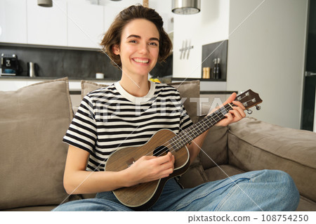 Beautiful girl playing ukulele on sofa, singing her favourite song and picking chords on small guitar, sitting in living room Beautiful girl playing ukulele on sofa, singing her favourite song and picking chords on small guitar, sitting in living room 108754250