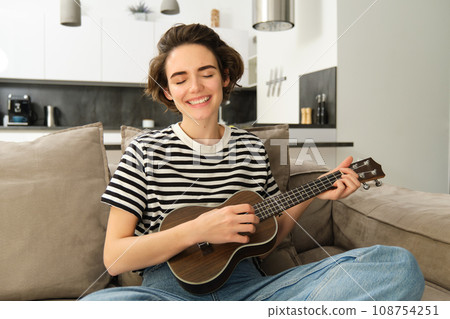 Young happy woman sitting on sofa and playing ukulele, singing and enjoying learning new musical instrument 108754251