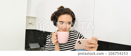 Smiling young woman watching tv series or video blog on smartphone app, drinking tea in the kitchen, looking at mobile phone with interest Smiling young woman watching tv series or video blog on smartphone app, drinking tea in the kitchen, looking at mobile phone with interest 108754253