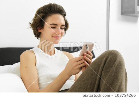 Cute young woman with mobile phone in hands, lying in bed in tank top, looking at smartphone with pleased smile Cute young woman with mobile phone in hands, lying in bed in tank top, looking at smartphone with pleased smile 108754260