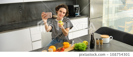 Portrait of beautiful young woman, food blogger recording video of her cooking in the kitchen, showing meal ingredients, preparing a meal on camera, using smartphone for live streaming 108754301