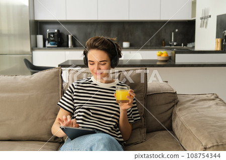 Close up portrait of modern girl, drinking orange juice, watching videos on digital tablet, sitting on sofa in living room Close up portrait of modern girl, drinking orange juice, watching videos on digital tablet, sitting on sofa in living room 108754344