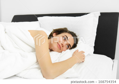 Portrait of young woman relaxing, lying in bed on soft white pillows, covered in linen comfortable sheets, smiling and looking outside with dreamy smile 108754354