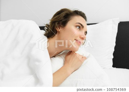 Close up portrait of beautiful young woman, lying in bed under white duvet, looks up at alarm clock, looking aside with relaxed smiling face 108754369