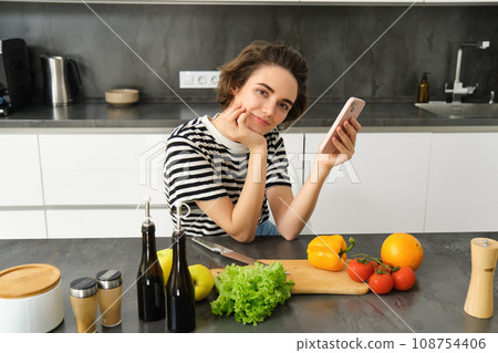 Portrait of cute brunette woman in the kitchen, cooking, searching recipe on smartphone, taking pictures for her food blog, cooking meal from vegetables, vegan salad Portrait of cute brunette woman in the kitchen, cooking, searching recipe on smartphone, taking pictures for her food blog, cooking meal from vegetables, vegan salad 108754406