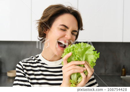 Portrait of happy and healthy young woman, following her diet, posing with lettuce leaf and smiling, cooking in the kitchen, vegetarian loves her vegetables Portrait of happy and healthy young woman, following her diet, posing with lettuce leaf and smiling, cooking in the kitchen, vegetarian loves her vegetables 108754428