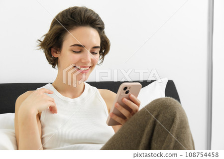 Beautiful woman lying in bed with smartphone, resting in her hotel suite, using mobile phone for food delivery, reading on cellphone Beautiful woman lying in bed with smartphone, resting in her hotel suite, using mobile phone for food delivery, reading on cellphone 108754458