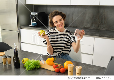 Smiling young woman, food blogger recording a video on smartphone with food, cooking and making selfie, taking pictures during meal preparation, sitting near vegetables and holding an apple 108754459