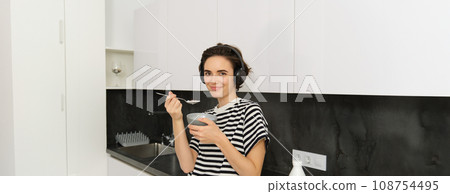 Cute young woman eats cereals with milk, holds spoon near mouth and smiles, listens music in wireless headphones, sits in kitchen Cute young woman eats cereals with milk, holds spoon near mouth and smiles, listens music in wireless headphones, sits in kitchen 108754495