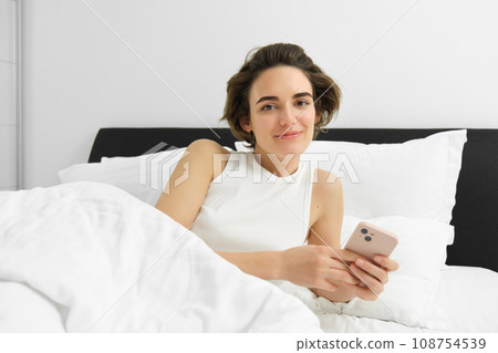 Portrait of young woman waking up in bed, using smartphone, turn off alarm on mobile phone, lying in her bedroom on white linen sheets Portrait of young woman waking up in bed, using smartphone, turn off alarm on mobile phone, lying in her bedroom on white linen sheets 108754539