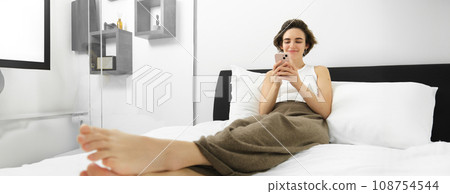 Photo of young woman resting in bedroom, lying in bed and using smartphone, looking at mobile phone and smiling, spending relaxing time at home Photo of young woman resting in bedroom, lying in bed and using smartphone, looking at mobile phone and smiling, spending relaxing time at home 108754544