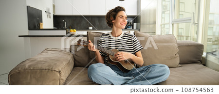 Portrait of girl resting in living room, playing ukulele, learns how to play new musical instrument, spending time relaxing at home Portrait of girl resting in living room, playing ukulele, learns how to play new musical instrument, spending time relaxing at home 108754563