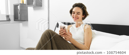 Portrait of young beautiful woman lying in bed, resting in bedroom, messaging, using mobile phone, holding smartphone and smiling, scrolling social media, spending comfort time at home 108754604
