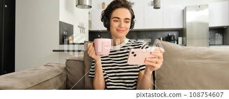 Portrait of woman in her living room, sitting on sofa and watching videos on mobile phone, wearing headphones, drinking tea or coffee in pink cup Portrait of woman in her living room, sitting on sofa and watching videos on mobile phone, wearing headphones, drinking tea or coffee in pink cup 108754607
