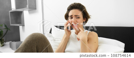 Portrait of woman in bed, answers phone call, looks worried and scared, hearing bad news, calling someone with concerned face 108754614