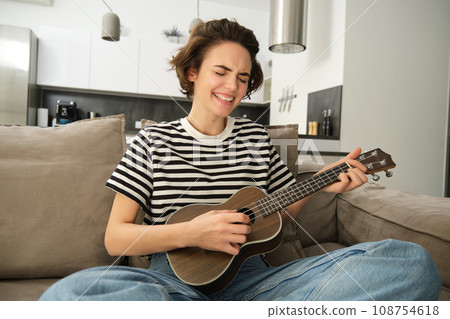 Portrait of young modern woman, student playing ukulele at home, sitting with small guitar, singing and feeling happy, sitting on sofa Portrait of young modern woman, student playing ukulele at home, sitting with small guitar, singing and feeling happy, sitting on sofa 108754618