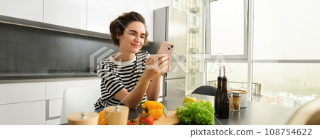 Portrait of beautiful smiling woman, cooking food at home, looking at smartphone, looking for healthy vegetarian recipe, standing near vegetables, making herself a salad 108754622