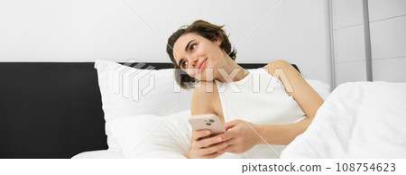 Portrait of woman lying in her bed, checking phone, sets up alarm clock to wake up in the morning Portrait of woman lying in her bed, checking phone, sets up alarm clock to wake up in the morning 108754623