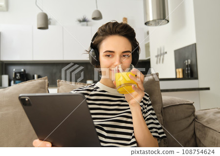 Close up portrait of modern girl, drinking orange juice, watching videos on digital tablet, sitting on sofa in living room Close up portrait of modern girl, drinking orange juice, watching videos on digital tablet, sitting on sofa in living room 108754624