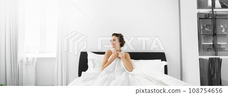 Portrait of beautiful, happy smiling woman, waking up in her bedroom, sitting in bed covered in linen sheets, looking outside 108754656