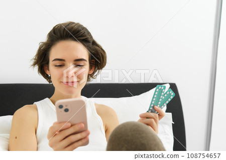 Portrait of woman in bed, holding mobile phone and pills, taking medication, searching for dosage and instruction for tablets, resting at home 108754657
