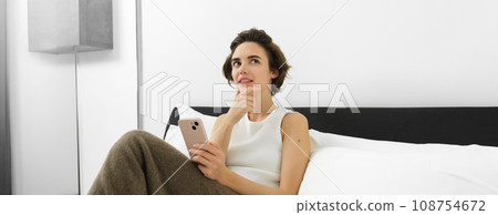 Portrait of beautiful woman with smartphone, makes thinking face, making decision, online shopping on mobile phone app while sitting in bed 108754672