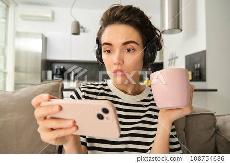 Portrait of woman with scared, concerned face watching video on smartphone, drinking tea, wearing headphones, sitting on sofa at home 108754686
