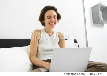 Modern woman resting at home, sitting on bed with laptop, looking at computer screen, laughing and smiling Modern woman resting at home, sitting on bed with laptop, looking at computer screen, laughing and smiling 108754740