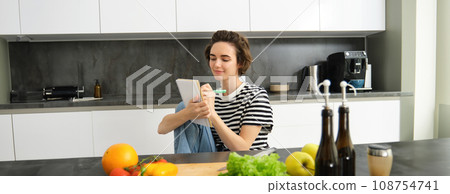Portrait of beautiful modern woman, girl cooking with vegetables, holding notebook, writing down recipe, making notes, dinner plans, creating meal list for week, sitting in kitchen Portrait of beautiful modern woman, girl cooking with vegetables, holding notebook, writing down recipe, making notes, dinner plans, creating meal list for week, sitting in kitchen 108754741
