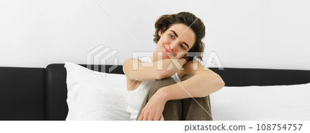 Young tender woman in her cosy pyjamas, sitting on bed in bedroom, smiling with sensual smile Young tender woman in her cosy pyjamas, sitting on bed in bedroom, smiling with sensual smile 108754757