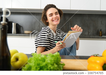 Portrait of young woman cooking, writing notes, grocery list in notebook, creating list of meals to cook through meal, sitting in kitchen near vegetables and chopping board Portrait of young woman cooking, writing notes, grocery list in notebook, creating list of meals to cook through meal, sitting in kitchen near vegetables and chopping board 108754785