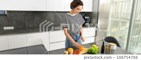 Portrait of modern young woman standing in the kitchen with vegetables, chopping them on counter, holding knife, cooking meal, vegetarian food, concept of healthy lifestyle an diet Portrait of modern young woman standing in the kitchen with vegetables, chopping them on counter, holding knife, cooking meal, vegetarian food, concept of healthy lifestyle an diet 108754788