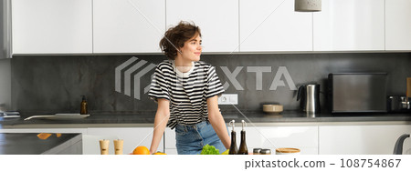 Portrait of young modern woman in the kitchen, leaning on counter with chopping board, making a salad with vegetables and dressings, cooking vegetarian meal Portrait of young modern woman in the kitchen, leaning on counter with chopping board, making a salad with vegetables and dressings, cooking vegetarian meal 108754867