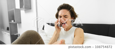 Young woman lying in bed, talking to friend over the phone, calling someone and complaining, gesturing with emotions, resting in bedroom Young woman lying in bed, talking to friend over the phone, calling someone and complaining, gesturing with emotions, resting in bedroom 108754872
