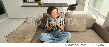 Portrait of happy brunette woman sitting on sofa and enjoying her breakfast, eating cereals, healthy granola in bowl, spending time in living room Portrait of happy brunette woman sitting on sofa and enjoying her breakfast, eating cereals, healthy granola in bowl, spending time in living room 108754873