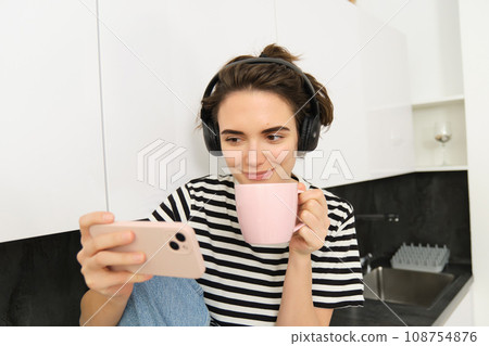 Smiling young woman watching tv series or video blog on smartphone app, drinking tea in the kitchen, looking at mobile phone with interest 108754876
