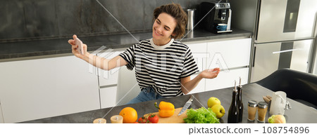 Portrait of beautiful young woman, food blogger recording video of her cooking in the kitchen, showing meal ingredients, preparing a meal on camera, using smartphone for live streaming Portrait of beautiful young woman, food blogger recording video of her cooking in the kitchen, showing meal ingredients, preparing a meal on camera, using smartphone for live streaming 108754896