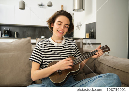 Beautiful young woman playing ukulele, singing and enjoying her musical instrument, spending time alone at home, relaxing in living room 108754901