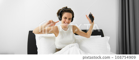 Portrait of happy young woman in bed, listening to music in wireless headphones, dancing in her bedroom, holding mobile phone in hand Portrait of happy young woman in bed, listening to music in wireless headphones, dancing in her bedroom, holding mobile phone in hand 108754909