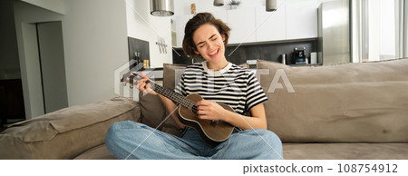Portrait of cheerful young woman playing her ukulele, singing and laughing, sitting in living room at home Portrait of cheerful young woman playing her ukulele, singing and laughing, sitting in living room at home 108754912