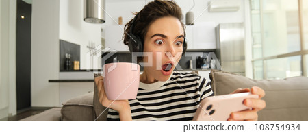 Portrait of woman binge watching her favourite tv show on smartphone, drinking tea and wearing headphones, gasping from shock, looking amazed at mobile screen 108754934