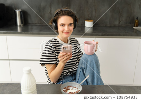 Modern female model smiling, using mobile phone, drinking tea and eating breakfast in kitchen 108754935